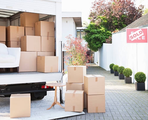 A moving truck full of packed boxes sits next to a white picket fence with a "sold" sign on it