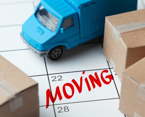 A toy moving truck sits on a paper desk calendar that says "moving" in red letters. There are cardboard boxes around the edge of the photo to imply moving your home.