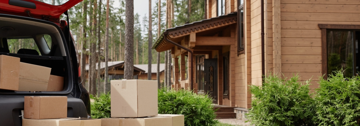 Background image of modern wooden house in woods with car and cardboard boxes in foreground, moving in living in nature