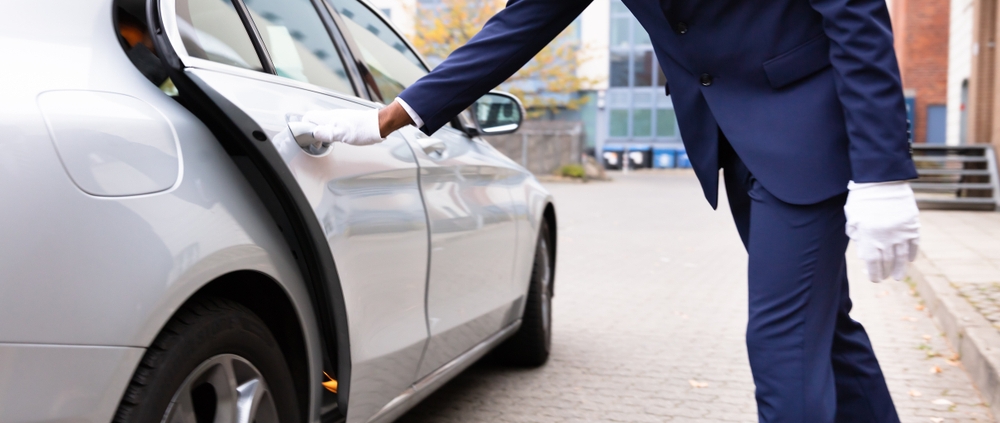 A man wearing a suit and clean white gloves opens a car door for a private car service client.