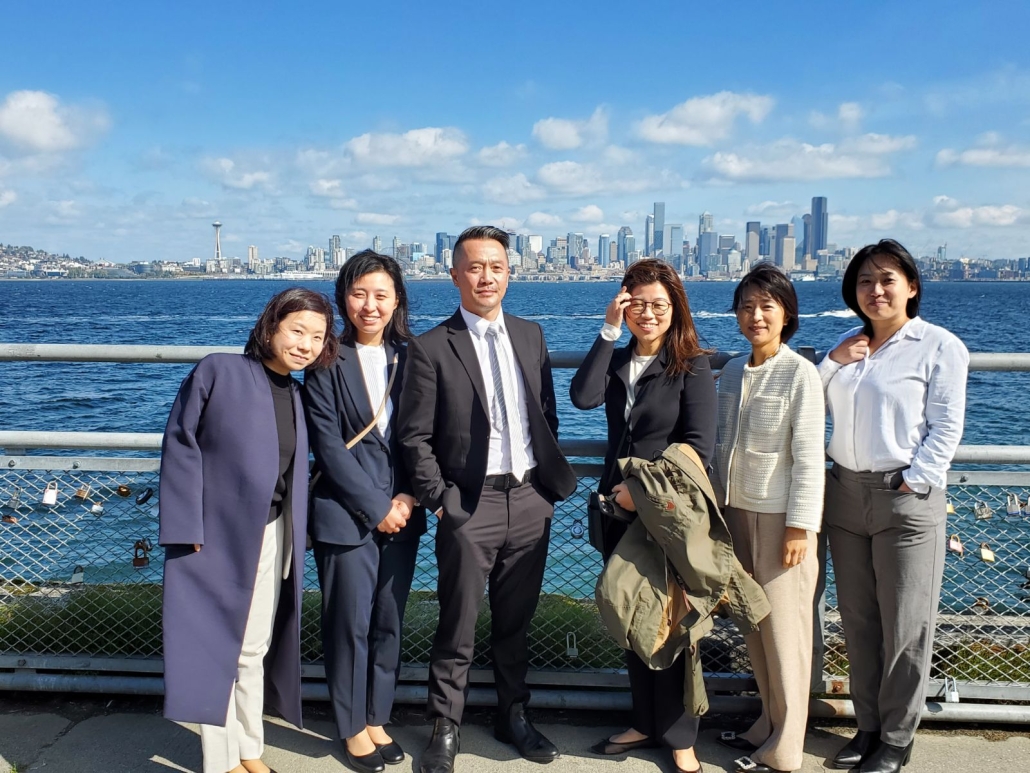 A man in a nice suit smiles, he is surrounded by 5 other people on a sunny day by the water in front of the Seattle skyline.